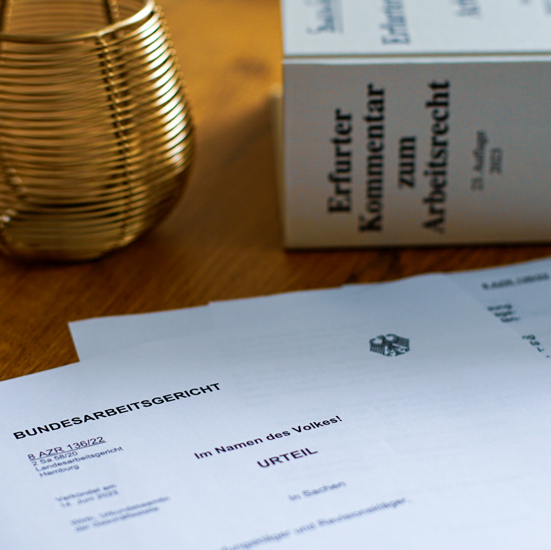Ein Entscheidung des Bundesarbeitsgerichts liegt auf einem Tisch. Im Hintergrund ist ein Buch mit dem Titel "Erfurter Kommentar zum Arbeitsrecht" zu sehen.